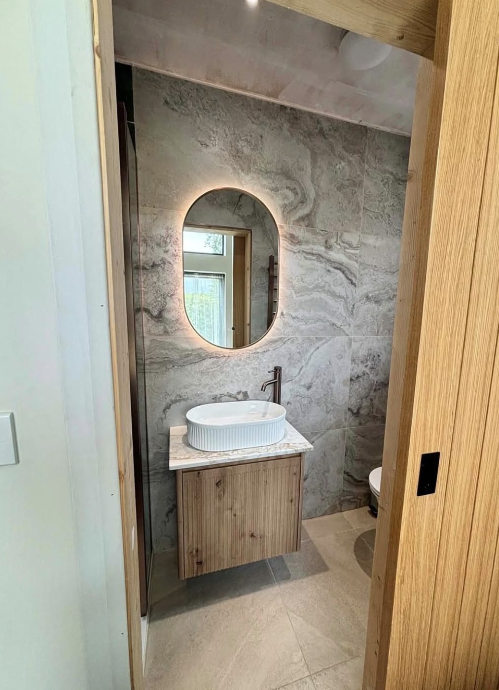 Cloakroom with a fluted basin on an oak vanity, oval backlit mirror and marble effect tiling