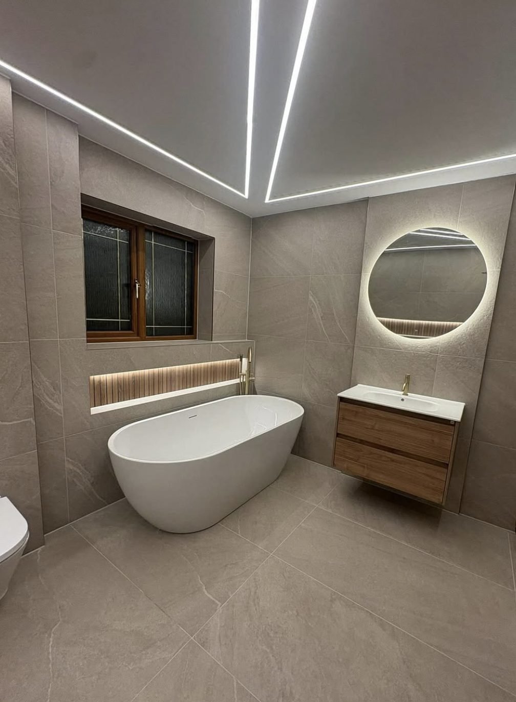 Freestanding bath in a stone effect tiled bathroom with LED ceiling strips and a round mirror