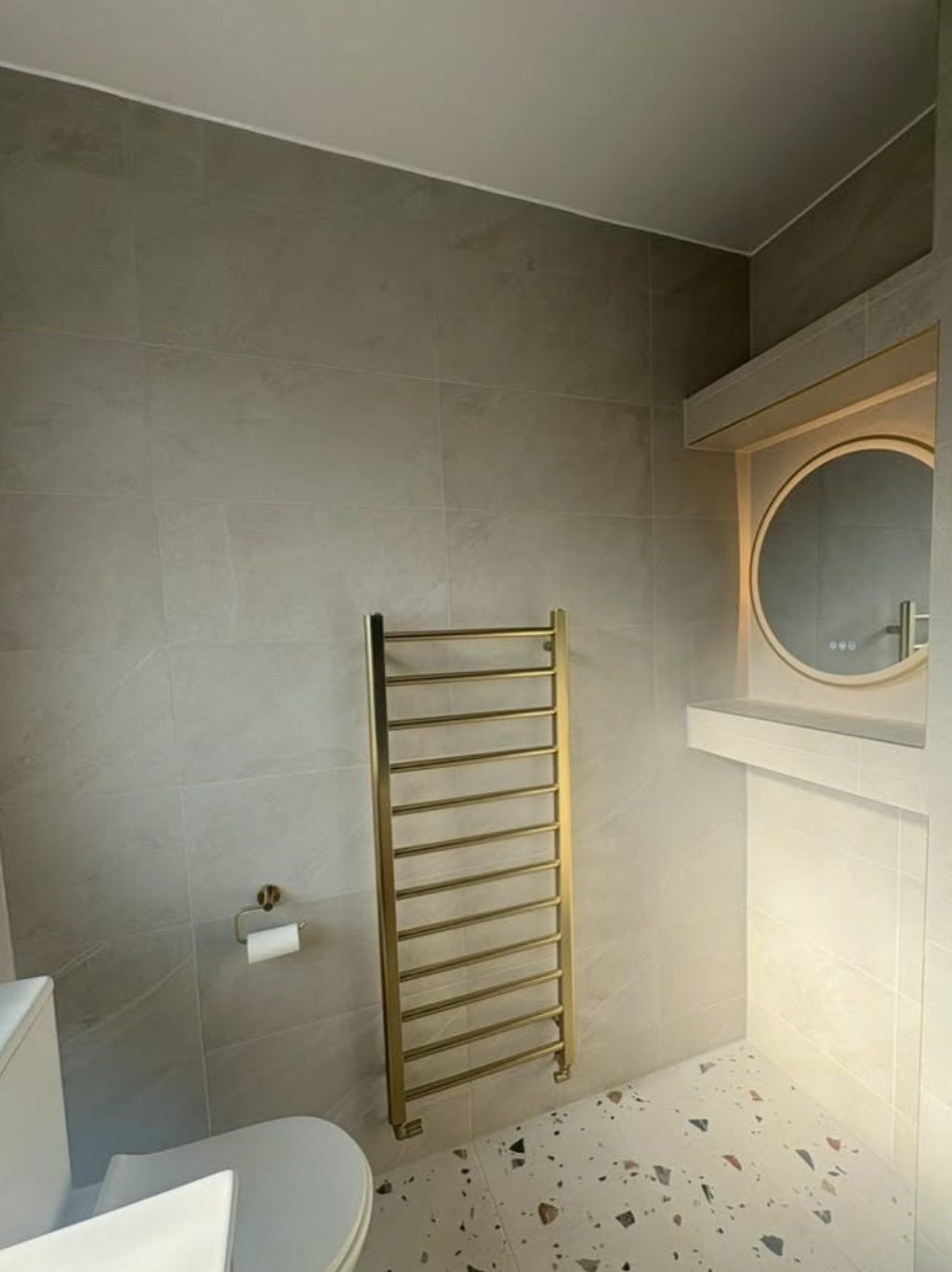 Grey tiled bathroom with a brushed gold ladder towel rail, round mirror and terrazzo floor